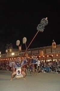 夜の祭り会場で毛槍を高々と持ち上げている男性の写真
