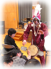 赤色の変わった服を着た人が座っている人たちの前で芸を披露している写真