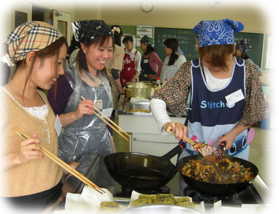 女性3人で1人が、木のしゃもじでフライパンの具材をかき混ぜ2人が菜箸を持ち眺めている写真