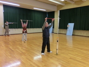 室内で三人の高齢者が棒を両手で持ち上げている写真